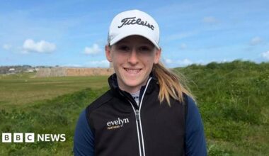 A man with short grey hair smiles into the camera. He's on a tee box at a golf course, with the sea and beach in the background, and a castle in the distance. He's wearing a dark blue jacket, branded with the golf club's logo.