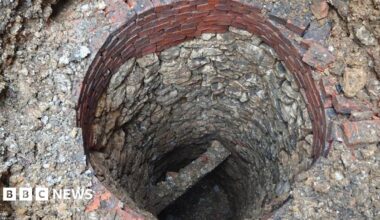 A circular well shaft lined with worn stones, looking down towards a stone cross beam and rubble at the base. Towards the top of the well are about eight layers of red bricks. More stones, rubble and bricks surround the well.
