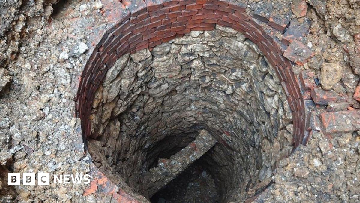 A circular well shaft lined with worn stones, looking down towards a stone cross beam and rubble at the base. Towards the top of the well are about eight layers of red bricks. More stones, rubble and bricks surround the well.