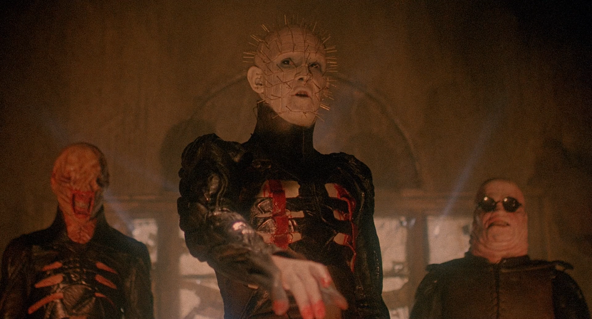 Three demonic monsters, the cenobites, stand in a room. They have distorted flesh.