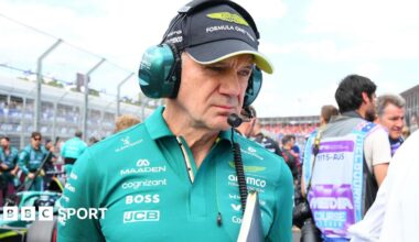 Adrian Newey pictured in an Aston Martin polo shirt at the Australian Grand Prix, with an Aston Martin car behind him to the right. Newey us holding a folder and wearing a headset