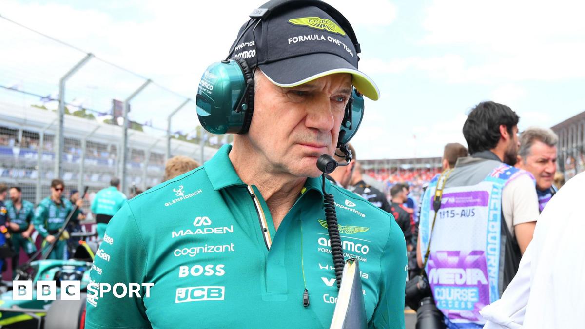 Adrian Newey pictured in an Aston Martin polo shirt at the Australian Grand Prix, with an Aston Martin car behind him to the right. Newey us holding a folder and wearing a headset