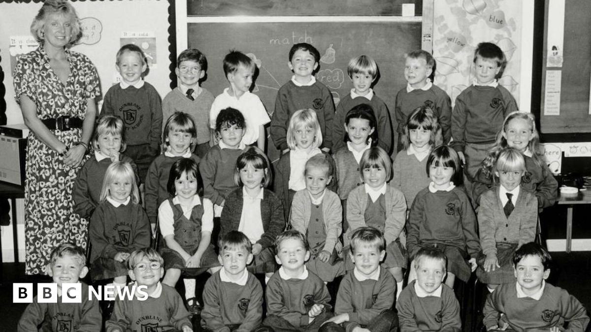 A black and white photo of Gwen Mayor, a blonde-haired woman in a patterned dress, and her 28 pupils arranged in four rows of seven in a classroom. The children are in school uniform.