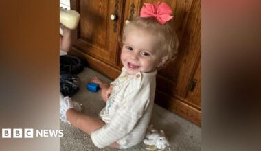 Viviana-Ray Butnaru - a young girl sitting on the floor, in front of a brown sideboard. She has turned her head to the left to smile at the camera. She has blonde hair with a pink bow in it and is wearing a white striped top.