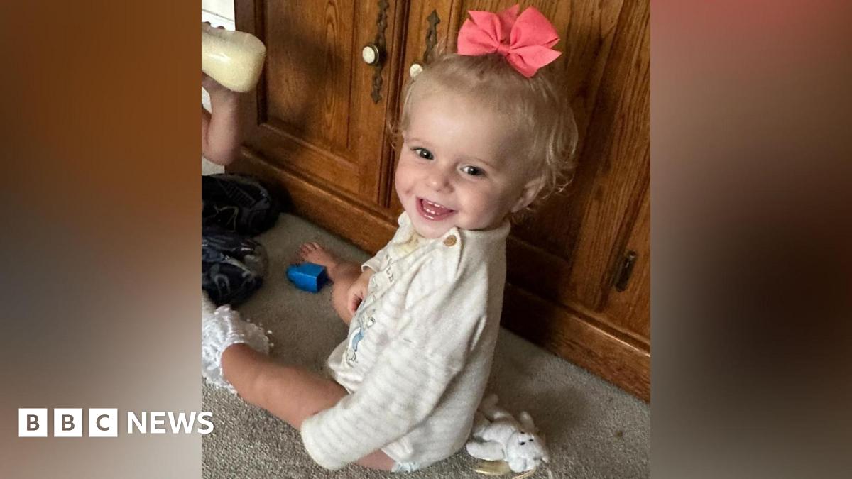Viviana-Ray Butnaru - a young girl sitting on the floor, in front of a brown sideboard. She has turned her head to the left to smile at the camera. She has blonde hair with a pink bow in it and is wearing a white striped top.