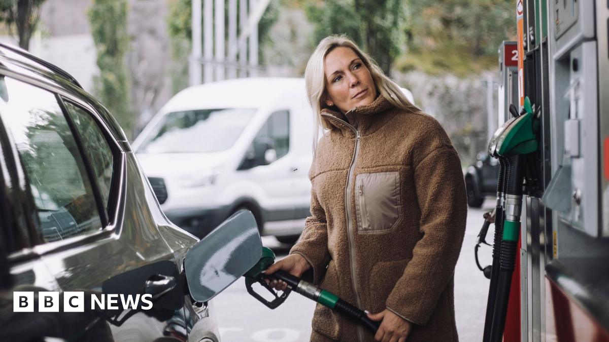 A stock photo of lady in a brown jacket filling up her car with fuel at a petrol station.