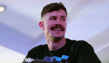 A man with short dark hair and a moustache with a black t-shirt in a white room