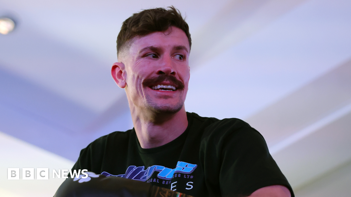 A man with short dark hair and a moustache with a black t-shirt in a white room