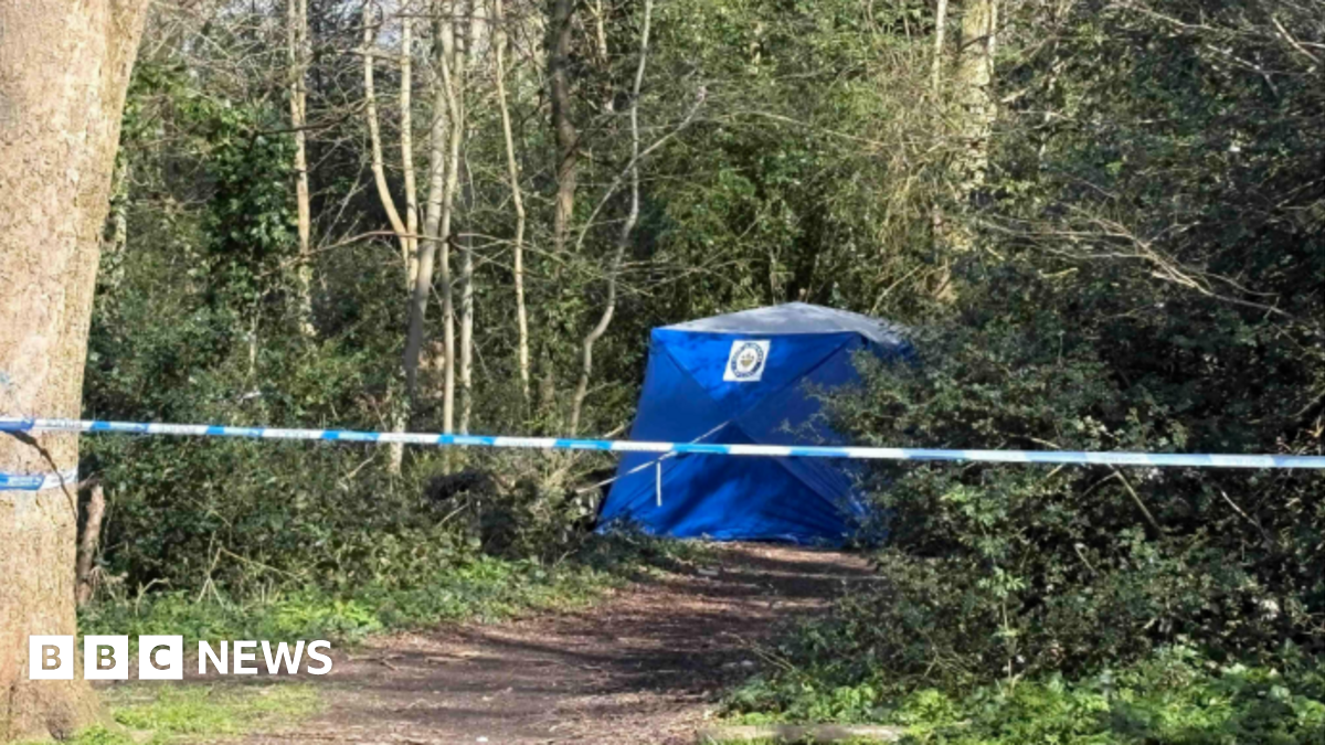 A police cordon attached to a tree in the middle of a wooded area. A blue police tent can be seen further down the footpath next to some trees.