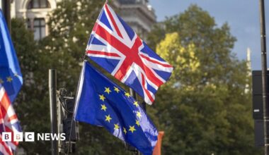 The flag of the United Kingdom flying above the EU flag. There are blurred trees behind the flags.