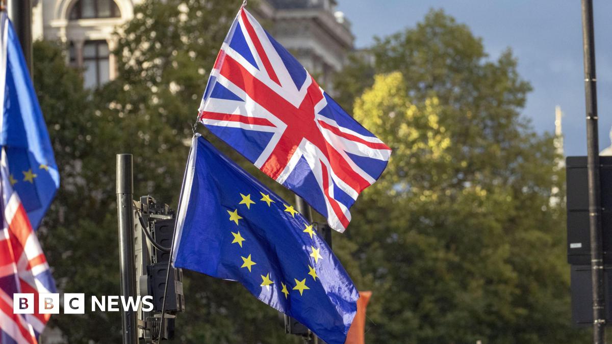 The flag of the United Kingdom flying above the EU flag. There are blurred trees behind the flags.