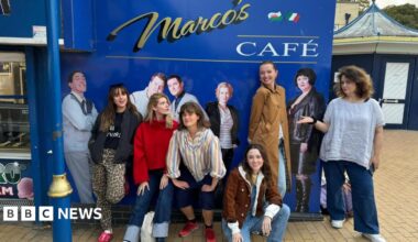 Five women are smiling while standing in front of the blue Marc's Cafe sign at Barry Island, with one woman kneeling on the floor. The sign includes photos of some of the cast of Gavin and Stacey, including Joanna Page as Stacey and Rob Bryden as Uncle Bryn. Ruth Jones is on the right, standing next to a photo of her as Ness.