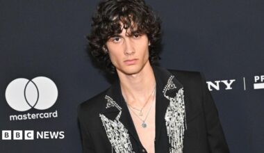 Sombr wearing a black jacket with ornate silver detailing stands before a dark event backdrop featuring Mastercard and Sony branding.