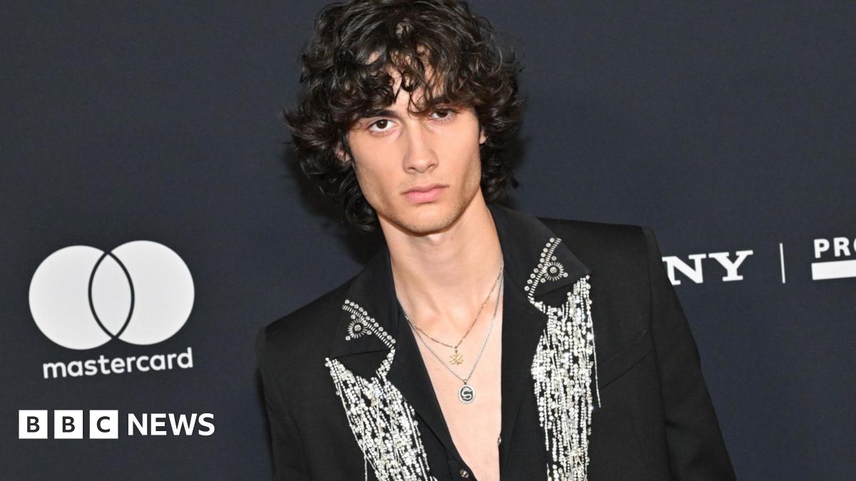 Sombr wearing a black jacket with ornate silver detailing stands before a dark event backdrop featuring Mastercard and Sony branding.