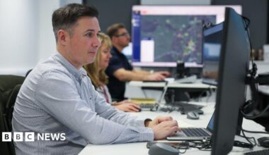 Sean Fryer has short brown hair and is wearing a chequered blue and white shirt. He is sitting at a desk working on a laptop and two colleagues, a woman with long brown hair and a man with short brown hair and glasses, are working at their desks in the background.