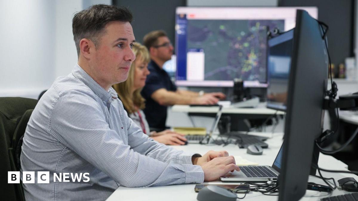 Sean Fryer has short brown hair and is wearing a chequered blue and white shirt. He is sitting at a desk working on a laptop and two colleagues, a woman with long brown hair and a man with short brown hair and glasses, are working at their desks in the background.