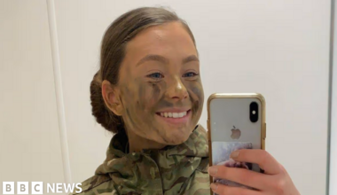 A mirror selfie of Jaysley Beck wearing a camoflauge jacket with green and brown face paint on her face. Her brown hair is tied up in a slick back low bun and she is smiling at the camera.