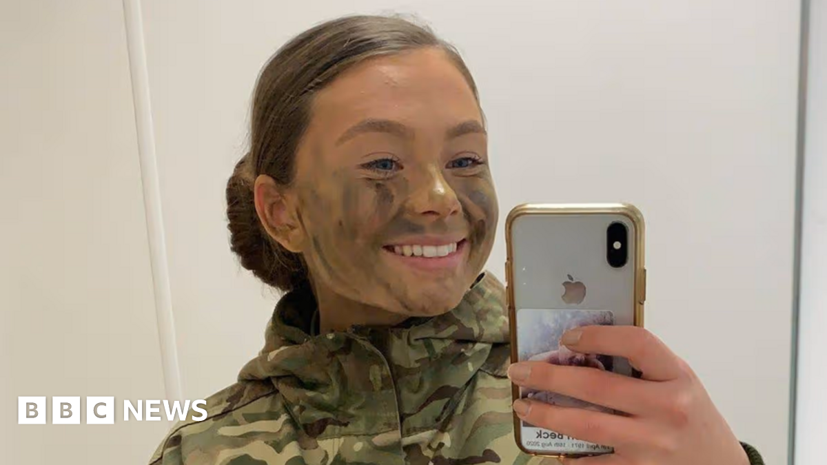 A mirror selfie of Jaysley Beck wearing a camoflauge jacket with green and brown face paint on her face. Her brown hair is tied up in a slick back low bun and she is smiling at the camera.