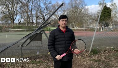 A man with dark hair and wearing a black jacket and trousers, red T-shirt and holding a tennis racket stands in front of the mangled fence with tennis courts behind it,