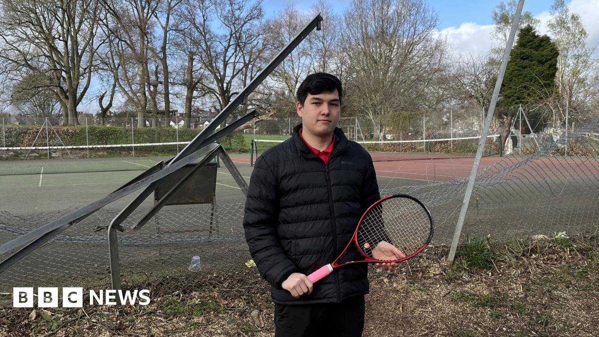 A man with dark hair and wearing a black jacket and trousers, red T-shirt and holding a tennis racket stands in front of the mangled fence with tennis courts behind it,