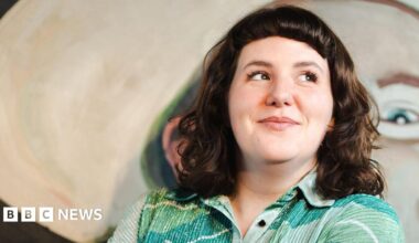 Amanda Dwyer standing in front of a large abstract painting with soft, rounded shapes and muted colours, wearing a patterned green shir