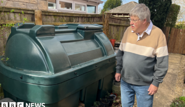 A man in a jumper stands next to a green oil tank