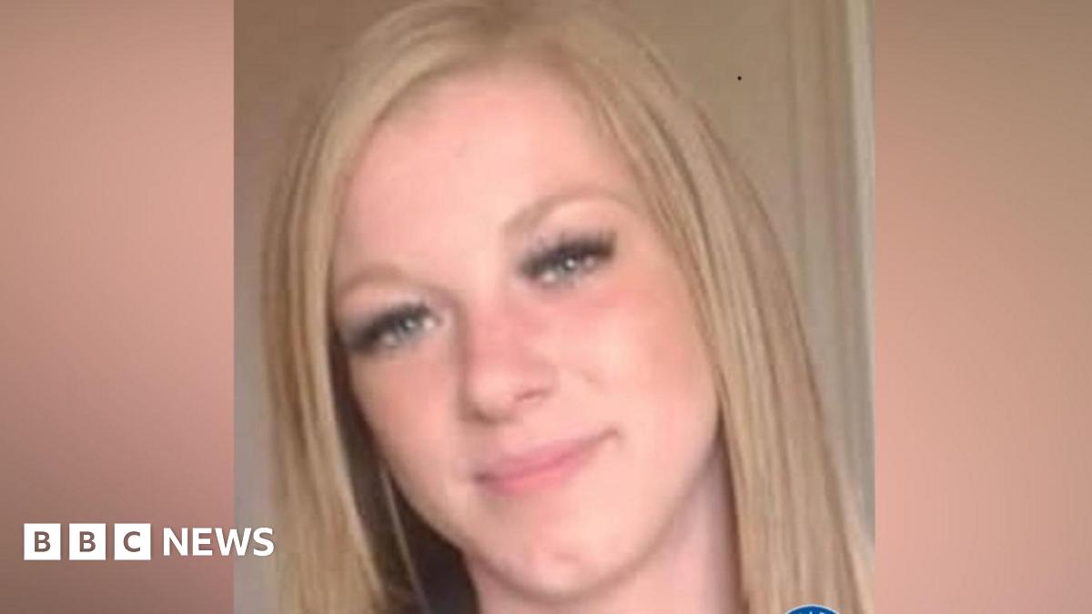A woman with pink long hair smiling. She is standing next to a wind