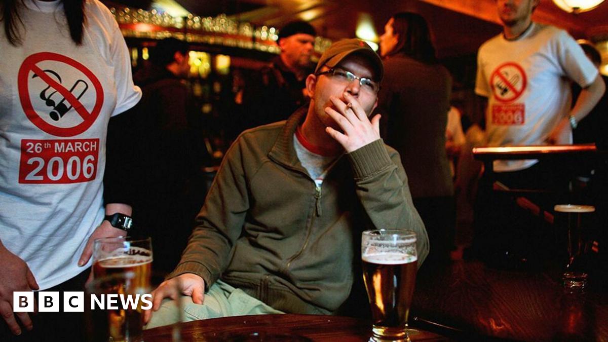 A man sitting at a table in a bar smokes a cigarette with a pint in front of him. Behind him, people are wearing t-shirts with a smoking ban symbol and the date of 26 March 2006