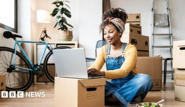 A young woman sitting on the floor in dungarees, smiling and wearing a head wrap to hold up her hair, she is working on a laptop which is propped up on a cardboard box - behind her there are a number of boxes and a ladder is propped against a wall, showing someone who has just moved