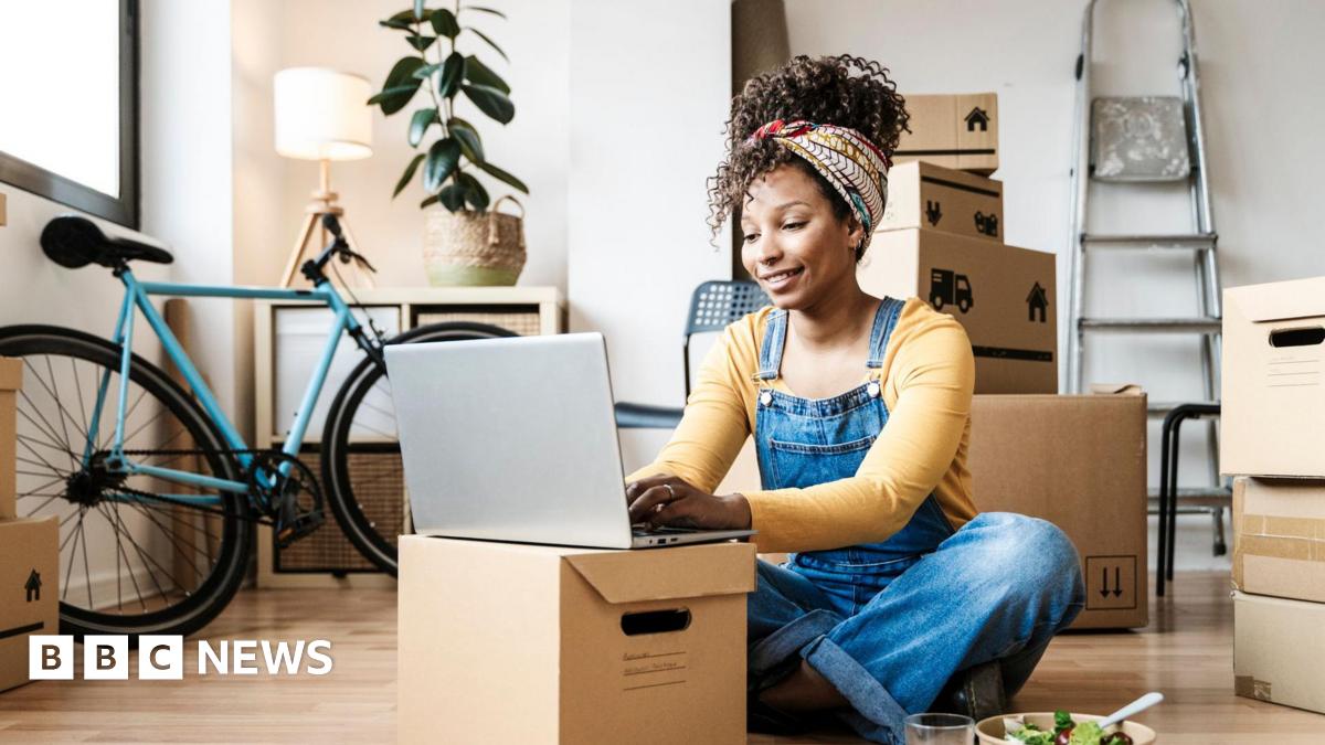 A young woman sitting on the floor in dungarees, smiling and wearing a head wrap to hold up her hair, she is working on a laptop which is propped up on a cardboard box - behind her there are a number of boxes and a ladder is propped against a wall, showing someone who has just moved