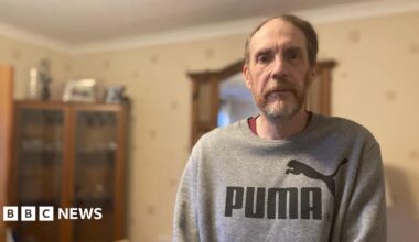 Gary Punter is wearing a grey puma sweatshirt and is standing in the back room of his rental property. He is looking at the camera and has beard. Behind him can be seen a corner, glass-fronted cupboard.
