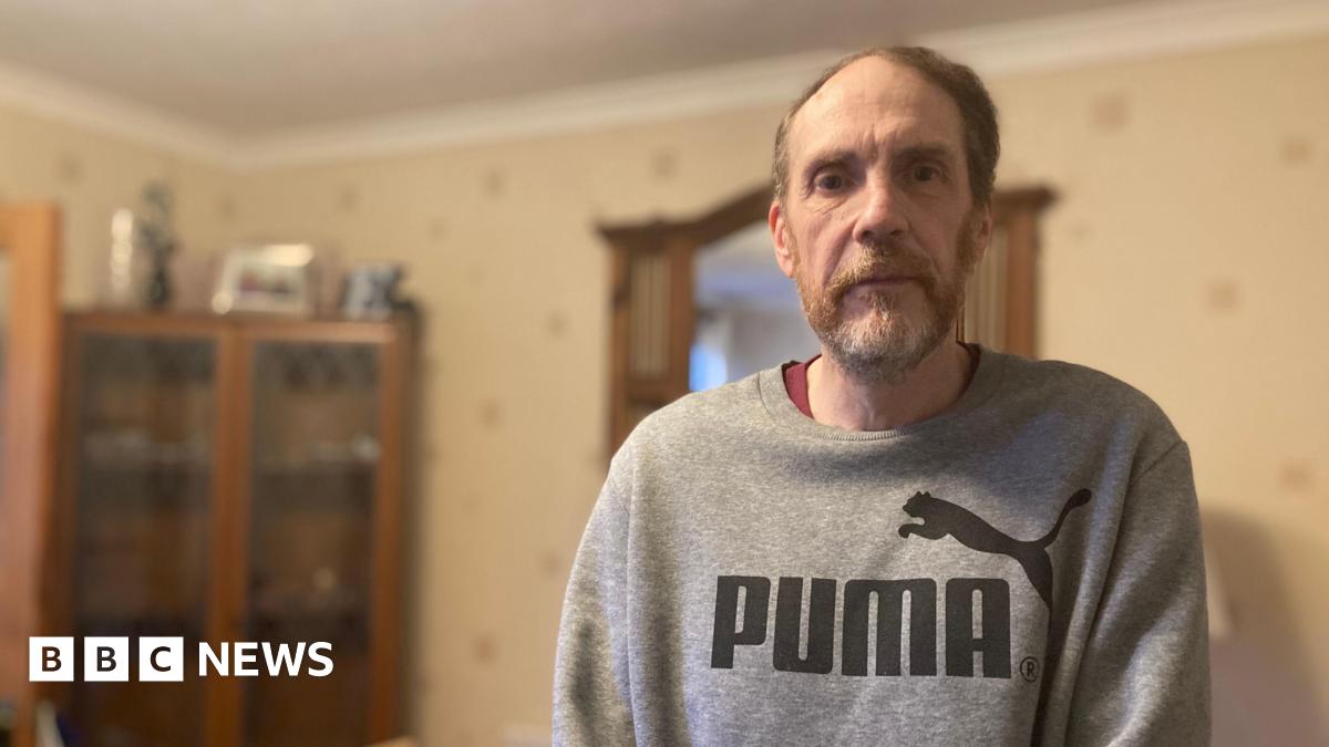 Gary Punter is wearing a grey puma sweatshirt and is standing in the back room of his rental property. He is looking at the camera and has beard. Behind him can be seen a corner, glass-fronted cupboard.