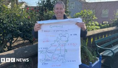 Man standing in a park in front of a bench. He is holding a map with the parishes in Guernsey written in Guernésiais.