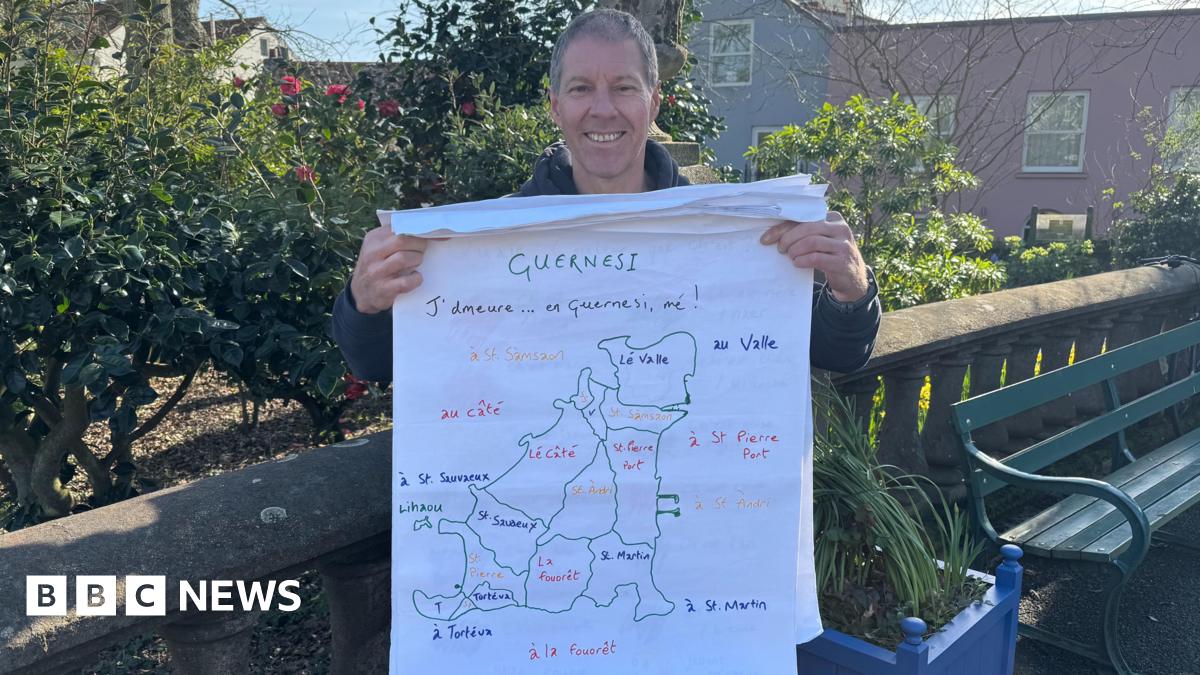 Man standing in a park in front of a bench. He is holding a map with the parishes in Guernsey written in Guernésiais.
