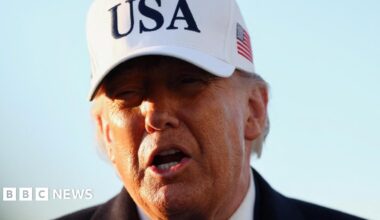 Trump pictured on 13 March. It is a close-up shot of his face in front of a blue sky. He wears a large white baseball cap with USA in large black letters on the front and a US flag on the side.