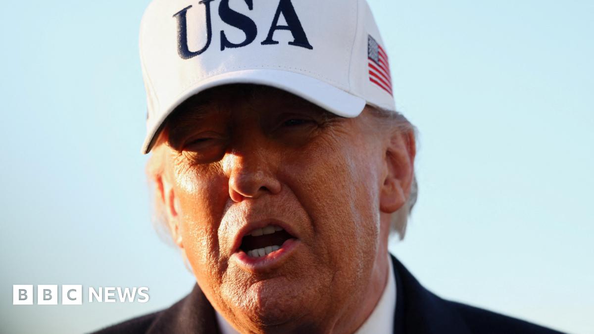 Trump pictured on 13 March. It is a close-up shot of his face in front of a blue sky. He wears a large white baseball cap with USA in large black letters on the front and a US flag on the side.