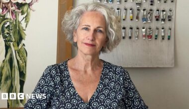 Marie Burke is sitting in front of a wall with paints hanging from nails. She is wearing a patterned shirt and has grey hair.