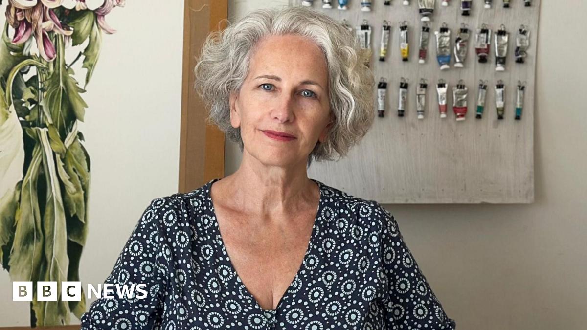 Marie Burke is sitting in front of a wall with paints hanging from nails. She is wearing a patterned shirt and has grey hair.