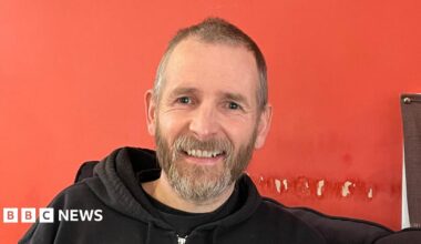A man with short brown and grey hair and a beard wearing a black hoodie sits against a red wall. He is smiling at the camera