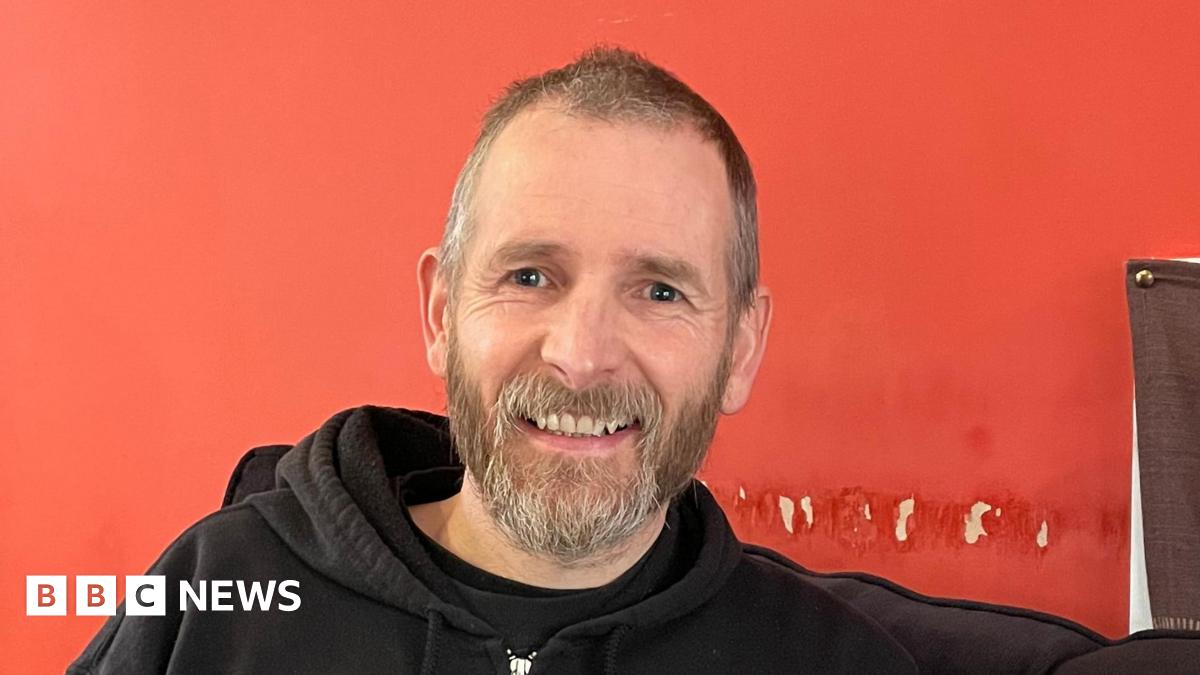 A man with short brown and grey hair and a beard wearing a black hoodie sits against a red wall. He is smiling at the camera
