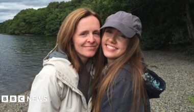 Beth, her mum, and Emily smile for the photo along a lake. Emily is wearing a grey cap and long brown hair whilst Beth has short gingery hair.
