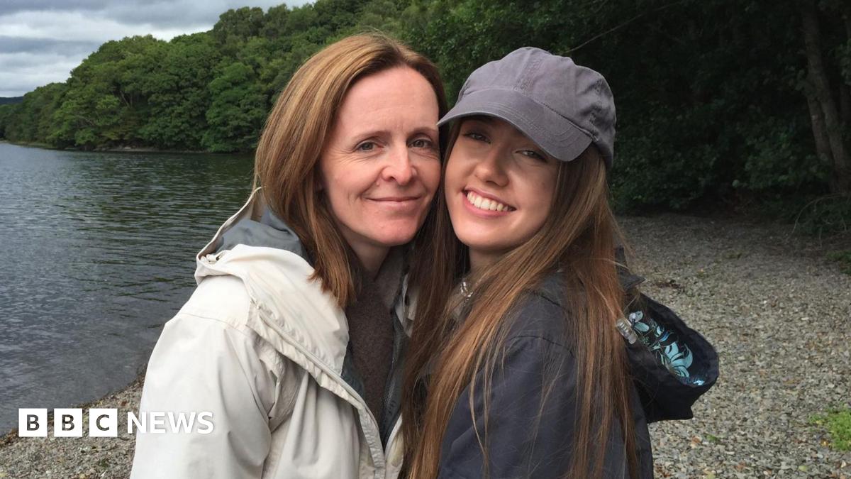 Beth, her mum, and Emily smile for the photo along a lake. Emily is wearing a grey cap and long brown hair whilst Beth has short gingery hair.