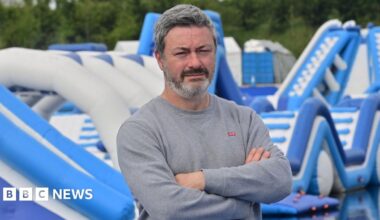 A man in a grey sweatshirt has his arms folded and is standing in front of blue and white water inflatables. He has greying short hair and black and grey beard.