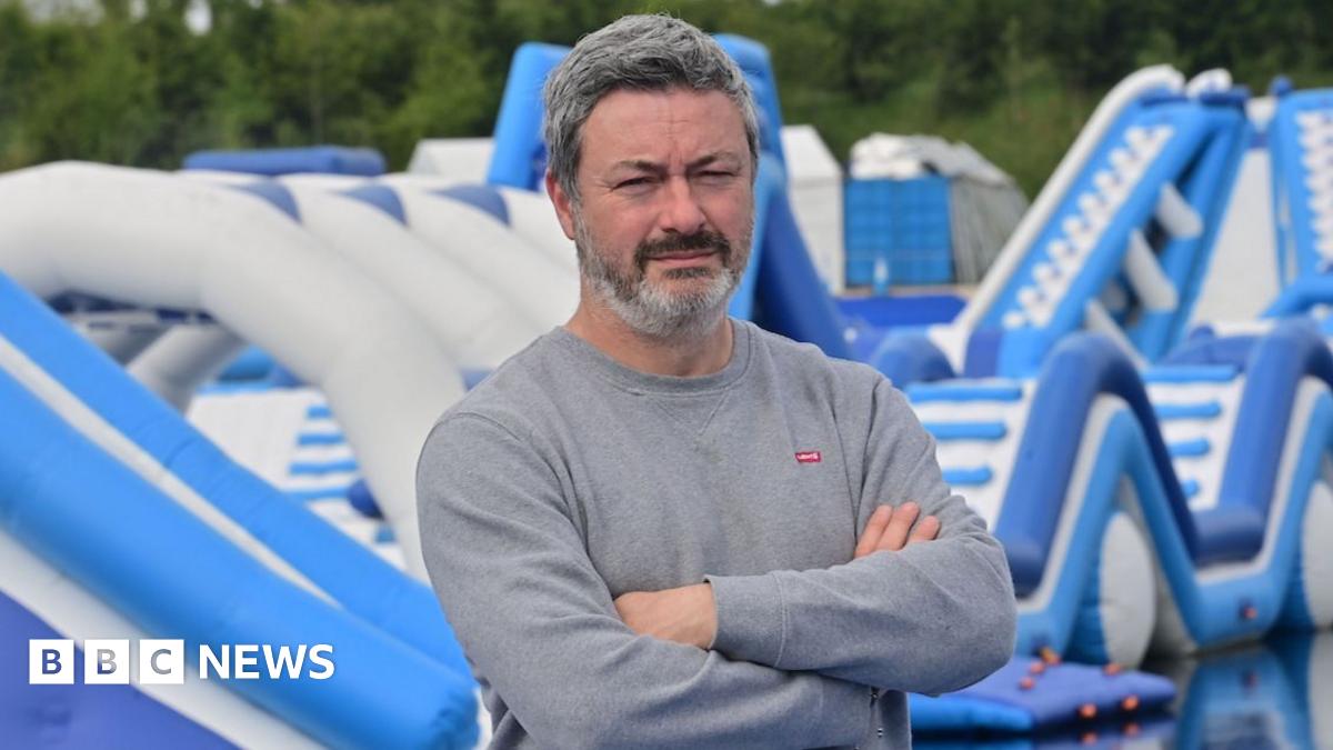 A man in a grey sweatshirt has his arms folded and is standing in front of blue and white water inflatables. He has greying short hair and black and grey beard.