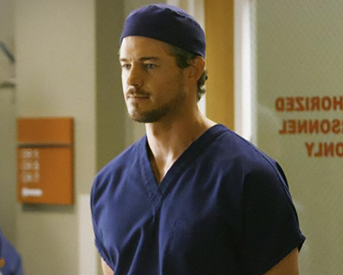 Eric Dane wearing navy blue scrubs and surgical cap standing in a hospital corridor scene.