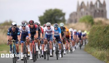 A group of cyclists, wearing colourful Lycra cycle along a paved, single track road, surrounded by hedgerows. In the background, Whitby Abbey, a ruined stone building, can be seen.