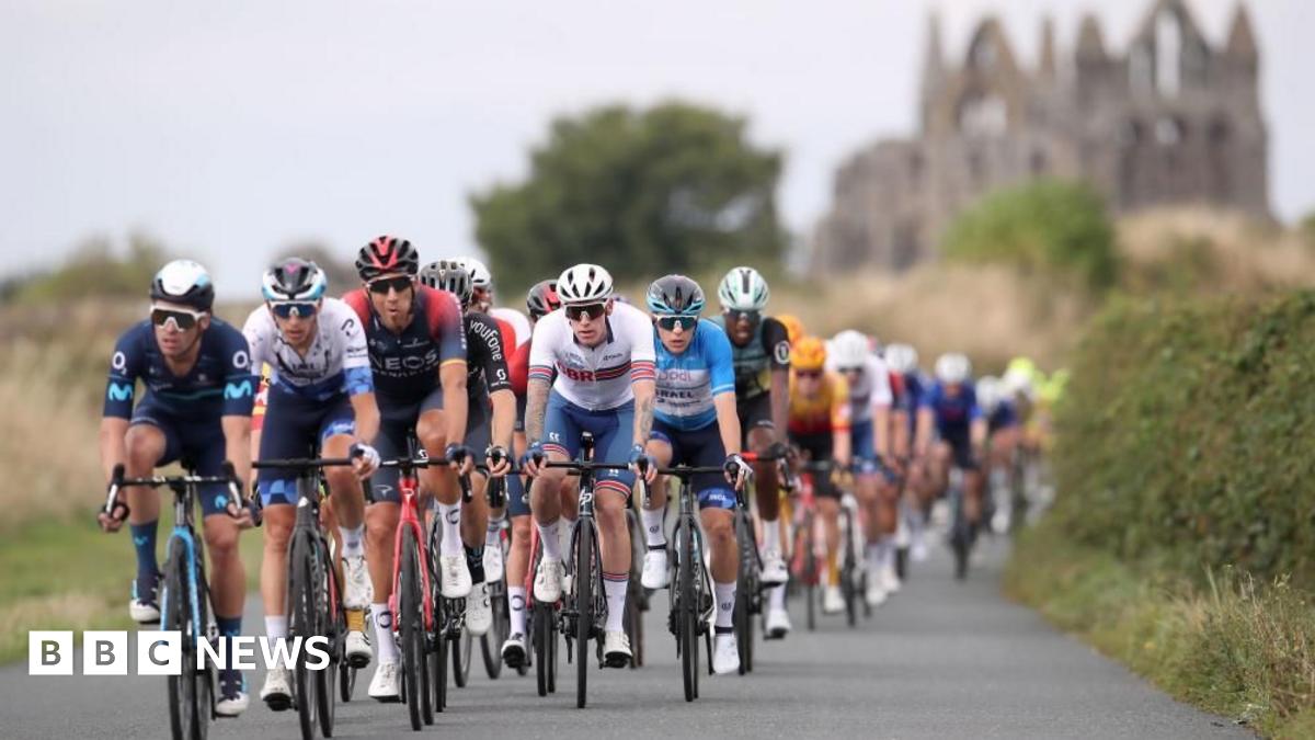 A group of cyclists, wearing colourful Lycra cycle along a paved, single track road, surrounded by hedgerows. In the background, Whitby Abbey, a ruined stone building, can be seen.