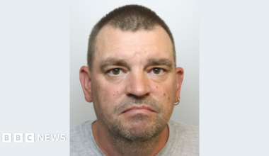A police mugshot of Matthew McKenzie, wearing a grey t-shirt and standing against a plain grey background. He has short brown hair, brown eyes, a silver hoop earring in his left ear and slight stubble. He is looking at the camera with a sad expression.