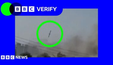 Young woman with red hair, wearing beige jumper holds smartphone in one hand and household bills in the other