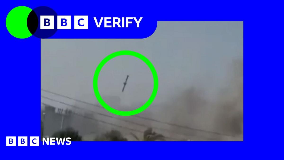 Young woman with red hair, wearing beige jumper holds smartphone in one hand and household bills in the other